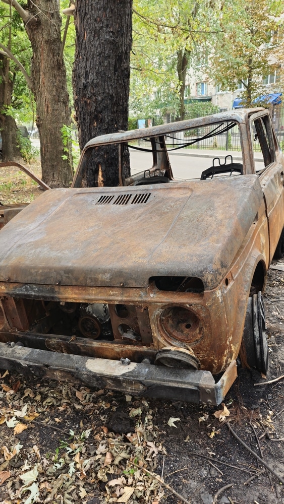 Épave de Lada Niva brûlée et rouillée : carcasse abandonnée Épave rouillée d'une Lada Niva calcinée abandonnée près d'un arbre dans un quartier urbain dévasté par le feu.
