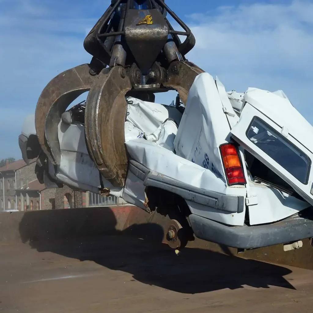 Pince de ferrailleur soulevant une fourgonnette blanche accidentée dans une casse automobile pour recyclage.