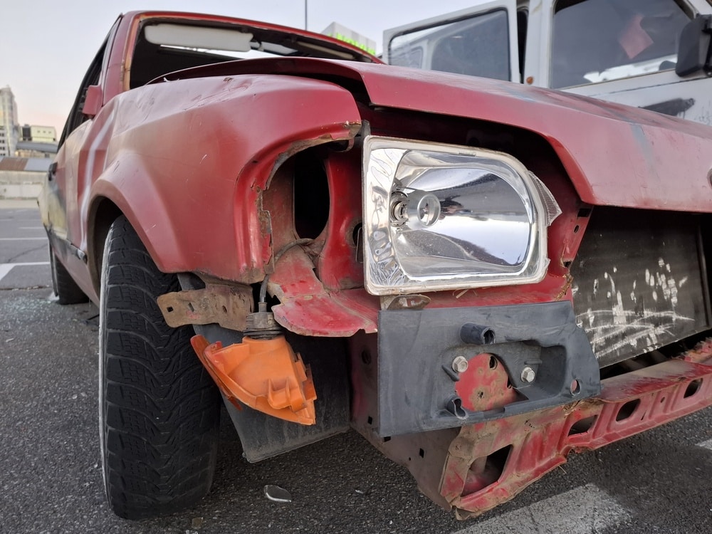 Voiture rouge accidentée : Gros plan carrosserie endommagée Voiture rouge accidentée avec phare brisé, clignotant pendant et carrosserie rouillée sur un parking urbain.